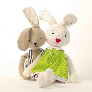 Può includere: Due giocattoli di peluche a forma di coniglio, uno marrone con strisce blu e bianche e l'altro bianco con un vestito a pois verdi.