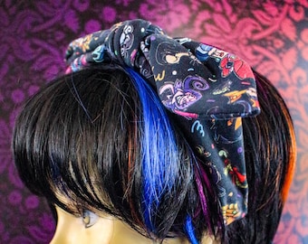 May include: A black fabric headband with a bow, patterned with colorful cartoon-like figures, is worn on a mannequin head with black hair and blue and orange streaks. The background is a dark purple floral pattern.