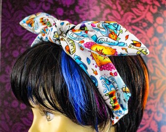 May include: A white headband with a colorful tattoo-style print featuring anchors, flowers, and other nautical elements. The headband is tied in a bow and worn on a mannequin with black hair and blue highlights. The background is a dark purple floral pattern.