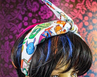 May include: A white headband with a colorful cartoon pattern featuring various characters. The headband is tied in a knot on top of the head. The mannequin has black hair with streaks of blue, orange, and pink.