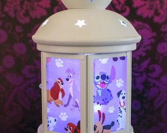 May include: A decorative, off-white lantern with star cutouts and a handle. The lantern features panels with cartoon dog characters and paw prints. The base is round and the background is a dark floral pattern.