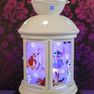 May include: A decorative, off-white lantern with star cutouts and a handle. The lantern features panels with cartoon dog characters and paw prints. The base is round and the background is a dark floral pattern.