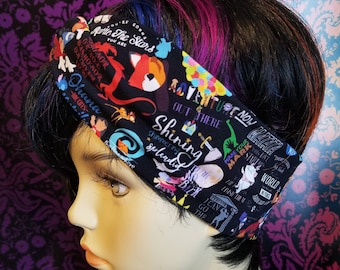 May include: Black fabric headband featuring colorful illustrations and text, including the words "Ohana," "Adventure," and "Shining." The headband is worn on a mannequin head with dark hair and streaks of pink and blue.