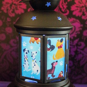 May include: Black metal lantern with blue star accents. The lantern features panels with cartoon dog illustrations on a light blue background. The base and top are round, and the lantern has a handle. The background is a dark floral pattern.