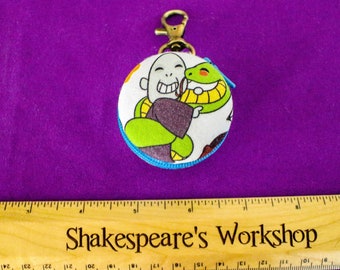 May include: A round, white fabric keychain with a cartoon illustration of a bald character hugging a green snake. The keychain has a blue trim and a gold-colored clasp. The background is a solid purple color.