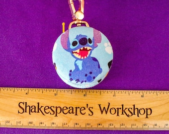 May include: A round, light blue keychain featuring a cartoon character with large eyes and a wide grin. The keychain has a gold-colored clasp and a matching ring. The text "Shakespeare's Workshop" is visible on a wooden ruler.