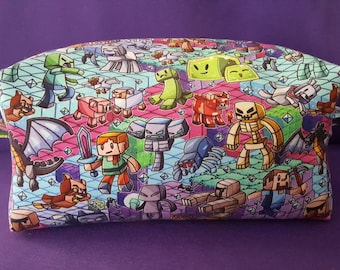 May include: A rectangular cosmetic bag with a colorful Minecraft-themed pattern. The design features various game characters and elements in a pixelated style, set against a light blue and pink background. The bag has a zipper closure and a small fabric loop on the side.