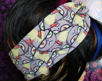 May include: A gray fabric headband featuring a repeating pattern of cartoon owl faces with yellow, red, and green accents. The headband is worn on a mannequin with black hair and colorful highlights.
