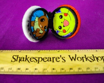 May include: A round, zippered plush case, open to reveal two cartoon character designs. One side features Eevee, the other Pikachu. The case is orange and white, with a black zipper. A wooden ruler with the text "Shakespeare's Workshop" is below.