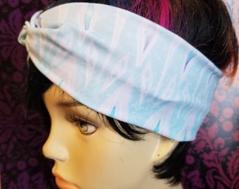 May include: A light blue and white patterned headband is worn on a mannequin head with black hair and pink and blue highlights. The headband has a knotted design and is made of a soft fabric. The background is a dark purple floral pattern.