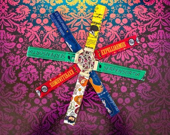 May include: A collection of colorful fabric keychains with various text and illustrations, including "Expelliarmus" and "Serpensortia". The keychains are attached to a silver metal ring and arranged in a starburst pattern against a patterned background.