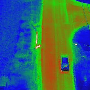 Può includere: Una vista aerea termica a infrarossi di un incrocio stradale che mostra le firme termiche. Un veicolo è visibile sulla strada in blu e verde, mentre la superficie stradale appare in arancione e giallo. Le linee elettriche attraversano l'inquadratura in diagonale.