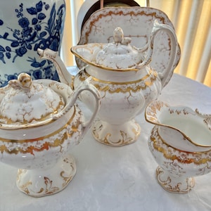 Könnte beinhalten: Ein Vintage-Teeservice mit weißem und goldenem Blumendesign. Das Set umfasst eine Teekanne, eine Zuckerdose und einen Sahnekännchen. Die Teekanne hat einen Deckel und einen gebogenen Ausguss. Die Zuckerdose hat einen Deckel und Griffe.