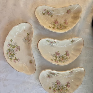 May include: Four antique porcelain dishes with a kidney bean shape. The dishes are off-white with a floral pattern in pink, green, and white. The rims are trimmed with gold.