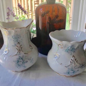 May include: Two vintage porcelain pitchers with a white background and blue and gold floral designs. The pitchers have a scalloped rim and a slightly rounded body.