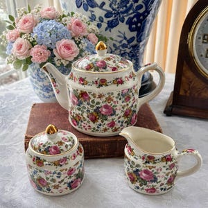 Puede incluir: Un juego de té floral vintage que incluye una tetera, un azucarero y una jarra para crema. Las piezas de porcelana blanca están adornadas con un colorido estampado floral de rosas rosas, flores azules y amarillas y hojas verdes. Cada pieza tiene detalles dorados.