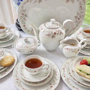 May include: A complete tea set with floral patterns. The set includes teacups, saucers, plates, a teapot, a creamer, a sugar bowl, and a serving platter. The tea cups are filled with tea, and a cookie and a sandwich are on the plates.