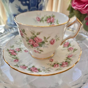 May include: A white teacup and saucer set with a floral rose pattern and gold trim. The teacup has a square handle and sits on a matching saucer. The set is designed for tea parties and special occasions.