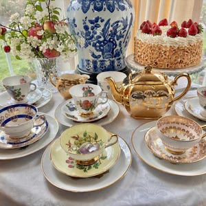 Könnte beinhalten: Eine Sammlung von Vintage-Tassen und Untertassen mit Goldrand, eine goldene Teekanne und ein Kuchen mit Erdbeeren. Eine große blau-weiße Vase und eine Blumenvase stehen ebenfalls auf dem Tisch.