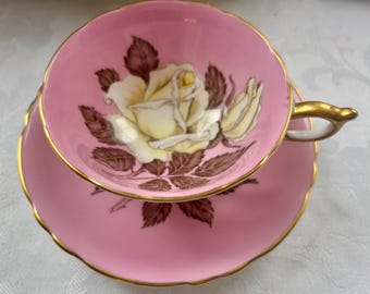 May include: A pink teacup and saucer set featuring a white and yellow rose design. The edges are trimmed with gold. The rose is accompanied by brown leaves. The teacup has a gold handle.