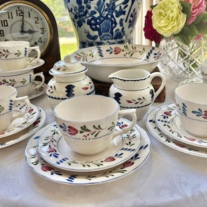 May include: A ceramic tea set featuring cream-colored pieces decorated with hand-painted blue and red floral patterns. The set includes multiple teacups with matching saucers, a lidded sugar bowl, a creamer pitcher, and a large serving bowl.