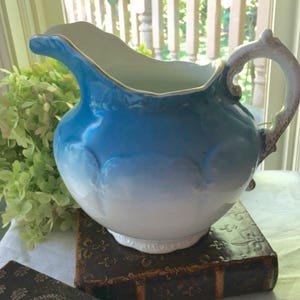 May include: A blue and white ceramic pitcher with a scalloped rim and a handle with a decorative design. The pitcher has a gradient of blue to white.