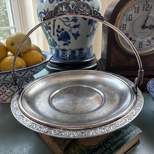 May include: A silver-toned serving tray with a decorative handle and ornate rim. The tray sits on a book and is surrounded by other decorative items, including a blue and white porcelain bowl filled with lemons and a vintage clock.