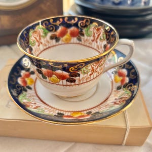May include: A vintage teacup and saucer set. The cup and saucer feature a navy blue border with gold trim and colorful floral designs. The teacup is resting on a small, light brown box.