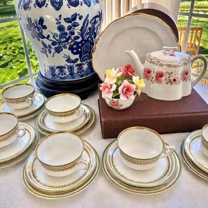 May include: A collection of vintage tea party items, including teacups, saucers, and a teapot. The white porcelain pieces have gold trim and a delicate floral design. A large blue and white vase is in the background, along with a small floral arrangement.