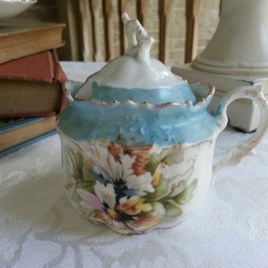 May include: A vintage porcelain sugar bowl with a lid, featuring a blue and white floral design with gold accents. The bowl has a scalloped edge and a delicate handle.