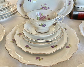 May include: A set of white porcelain dinnerware with a delicate floral pattern featuring purple violets. The set includes a teacup, saucer, and plates of varying sizes.