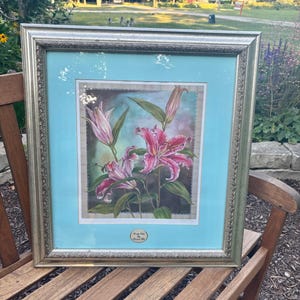 May include: Framed floral art featuring pink and white lilies with green leaves against a light blue background. The artwork is in a silver-toned frame and is displayed on a wooden bench outdoors.