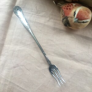May include: A silver, ornate, three-pronged fork with a decorative handle. The fork is on a light brown fabric background.