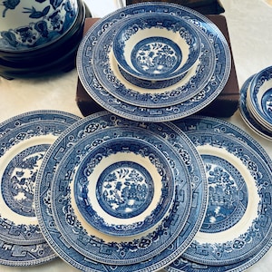 Puede incluir: Una colección de vajilla de cerámica azul y blanca, que incluye platos y cuencos, con un diseño clásico de Willow. El juego incluye varios tamaños de platos y cuencos, perfectos para servir una comida. Los platos están colocados sobre una superficie blanca.