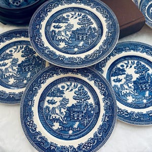 May include: A collection of blue and white ceramic plates with a classic Willow pattern design. The plates feature a central landscape scene surrounded by a decorative border. Several plates are visible, along with a bowl and a stack of smaller plates.