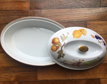 May include: White oval ceramic serving dish with a lid. The lid has a design of pears, lemons, asparagus, and blueberries on a white background.