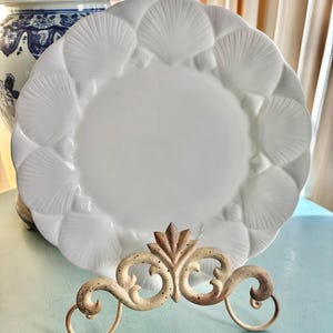 Könnte beinhalten: Ein weißer Keramikteller mit einem Muscheldesign am Rand. Der Teller wird auf einem dekorativen, verzierten Metallständer präsentiert. Im Hintergrund ist eine blau-weiße Porzellankanne zu sehen.