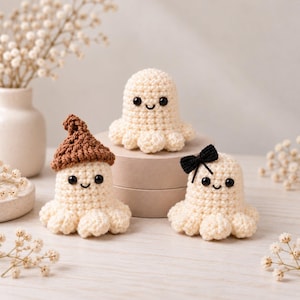 Puede incluir: Tres pequeñas figuras de fantasmas de ganchillo color crema con ojos de cuentas negras y sonrisas bordadas. Un fantasma lleva un gorro de ganchillo marrón, otro tiene un pequeño lazo negro y el tercero es sencillo. Base redondeada.