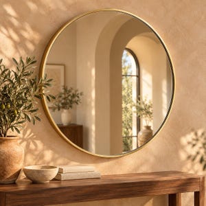 May include: A large circular wall mirror featuring a thin, smooth gold-toned metal frame. The reflective glass surface provides a clear view, capturing the surrounding interior space, including an arched doorway and natural light reflections.