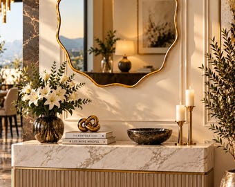 May include: A scalloped gold-framed wall mirror hangs above a rectangular console table with a white marble top and a ribbed beige base. The table holds a glass vase with white lilies, a gold knot sculpture, two books, and a dark marble bowl.