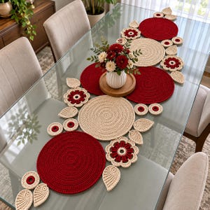 Könnte beinhalten: Ein Häkel-Tischläufer mit abwechselnd großen, kreisförmigen Matten in Dunkelrot und Beige, akzentuiert durch kleinere blumenförmige Motive und blattförmige Teile in passenden Farben, zentriert mit einer weißen Vase voller roter und weißer Blumen.