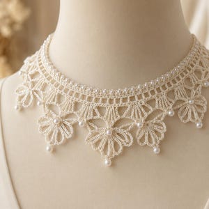 May include: A cream-coloured crochet choker necklace featuring a delicate floral pattern with intricate thread work. The design is accented with small, round white pearls embedded along the top edge and at the centre of each flower petal motif.