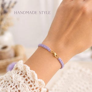 Puede incluir: Una pulsera de ganchillo color lavanda con un cierre de mosquetón dorado. La delicada banda de hilo tejido envuelve la muñeca, mostrando un diseño sencillo y texturizado. El texto HANDMADE STYLE es visible en la esquina superior izquierda.