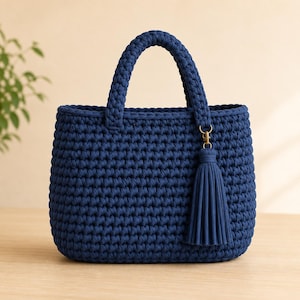 Puede incluir: Un bolso de mano azul marino profundo elaborado con un punto de ganchillo grueso y texturizado. El bolso cuenta con dos asas redondeadas resistentes y una borla azul marino a juego sujeta a un asa con un pequeño cierre de metal.