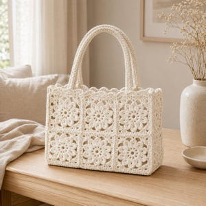 May include: A cream-coloured tote bag constructed from six square crochet panels featuring a central floral motif. The bag includes two matching crochet handles and a scalloped top edge, showcasing a consistent yarn texture throughout the design.
