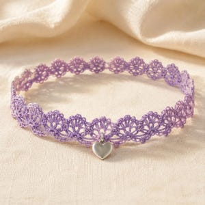 May include: A lavender lace choker necklace featuring a delicate scalloped edge pattern and a single, smooth, silver-toned heart-shaped charm suspended from the centre of the band. The lace is crafted with a fine, intricate thread design.