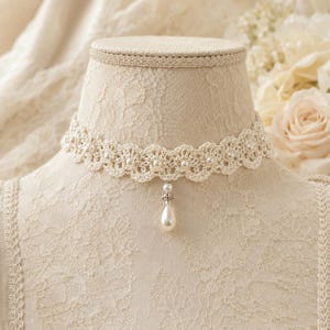 May include: A cream-colored crochet choker necklace featuring a scalloped edge design. The lace band is embellished with small, round white pearls integrated into the pattern, and a single teardrop-shaped pearl pendant hangs from the center.