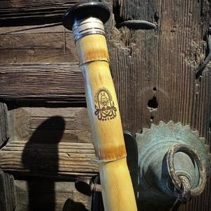 May include: A light-coloured bamboo flute featuring a black circular cap at the top, silver-toned metal rings, and a dark brown printed emblem on the body. The instrument has a smooth, cylindrical surface with visible natural bamboo nodes.