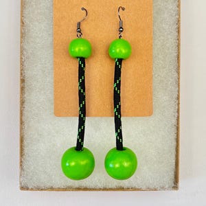 May include: A pair of drop earrings featuring a black cord with a green pattern, each accented by a small green bead at the top and a larger green sphere at the bottom, suspended from dark metal hooks on a rectangular brown display card.