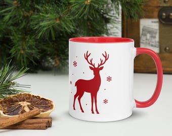 May include: A white ceramic mug with a red interior and a red handle, featuring a red silhouette of a standing reindeer surrounded by small red snowflake icons on the front surface of the mug.